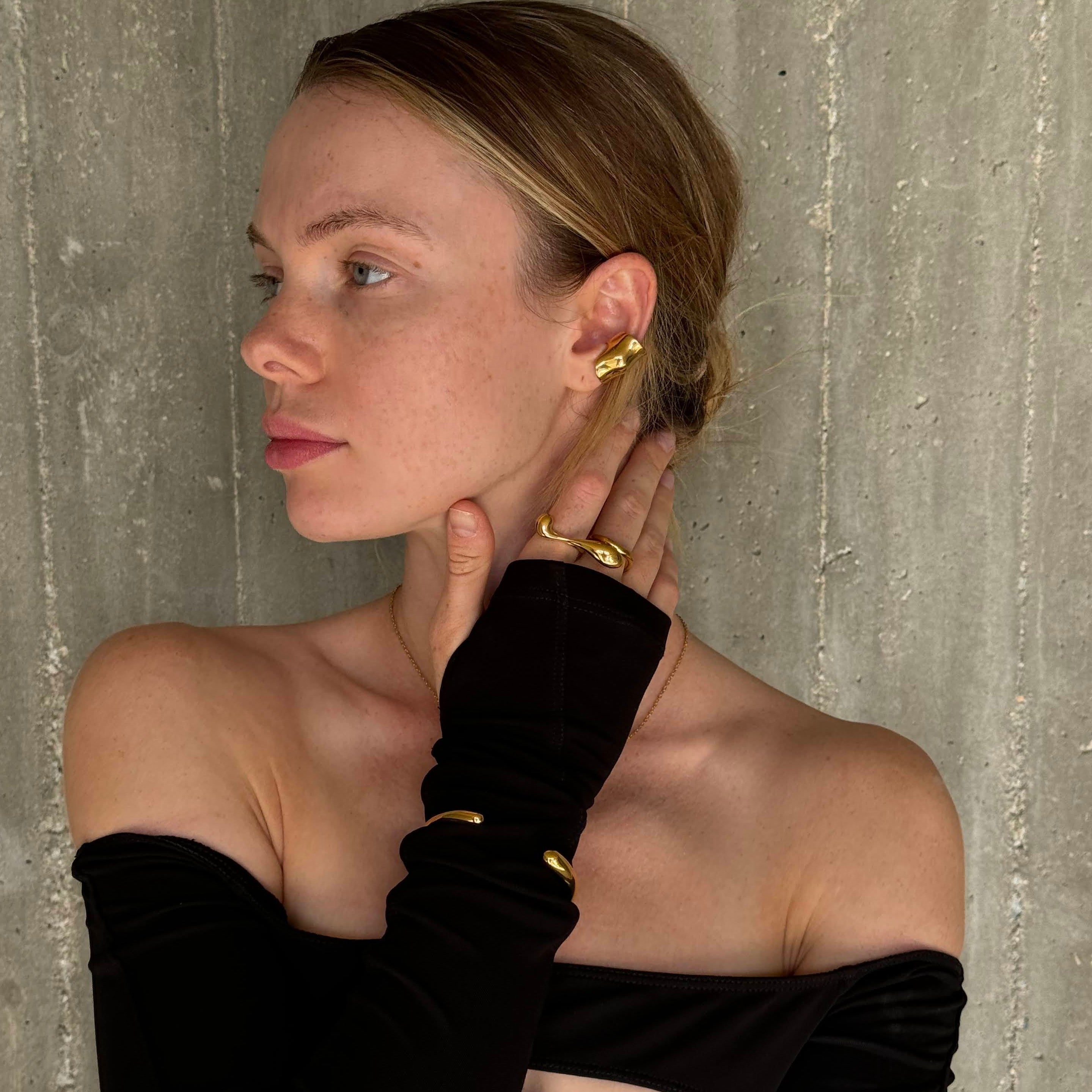 Model in a black off-shoulder top wearing the long drop-shaped ring in PVD-coated 18k gold-plated stainless steel with her hand to her neck and matching gold earrings