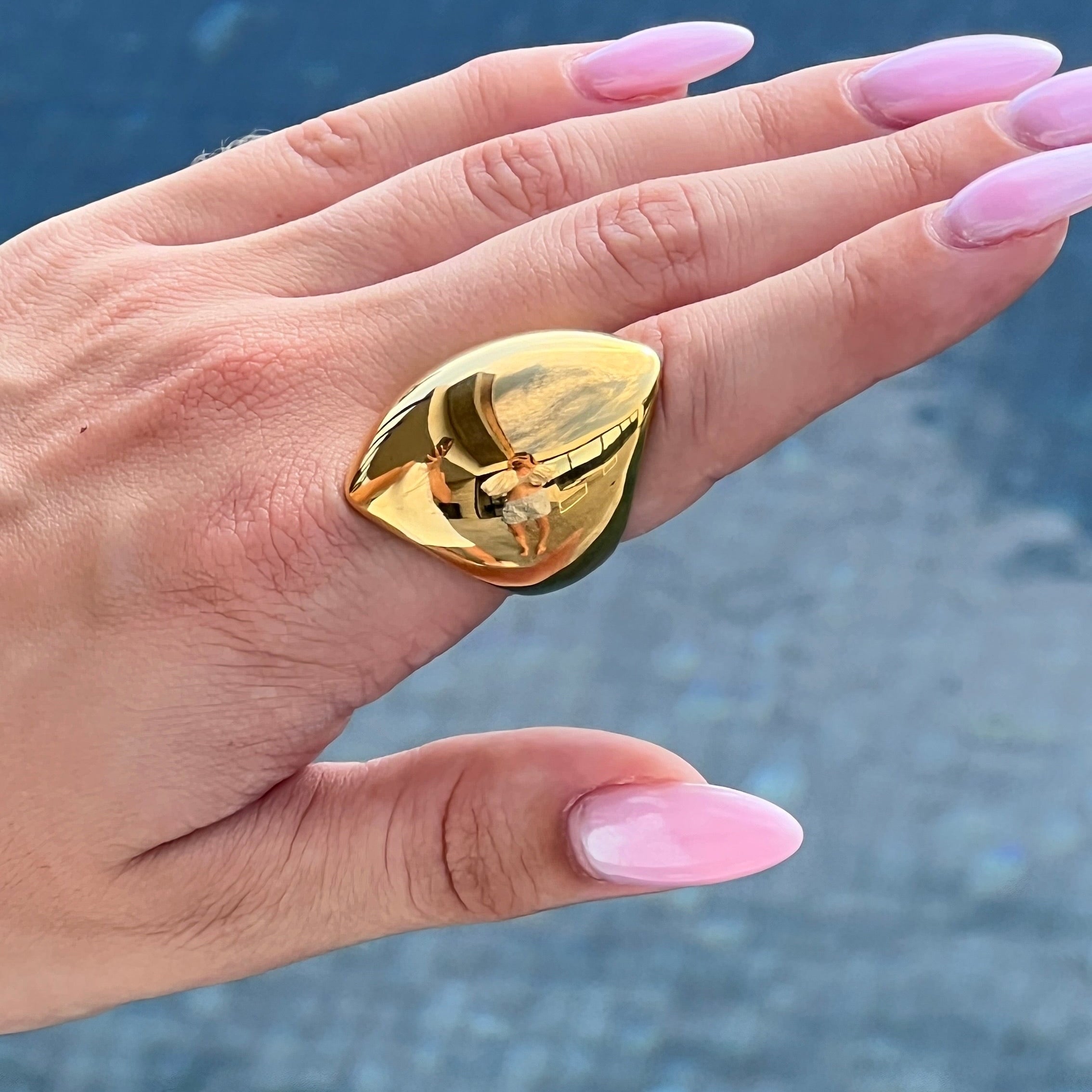 Hand with pink manicure wearing the angular ring in PVD-coated 18k gold-plated stainless steel against a blue background