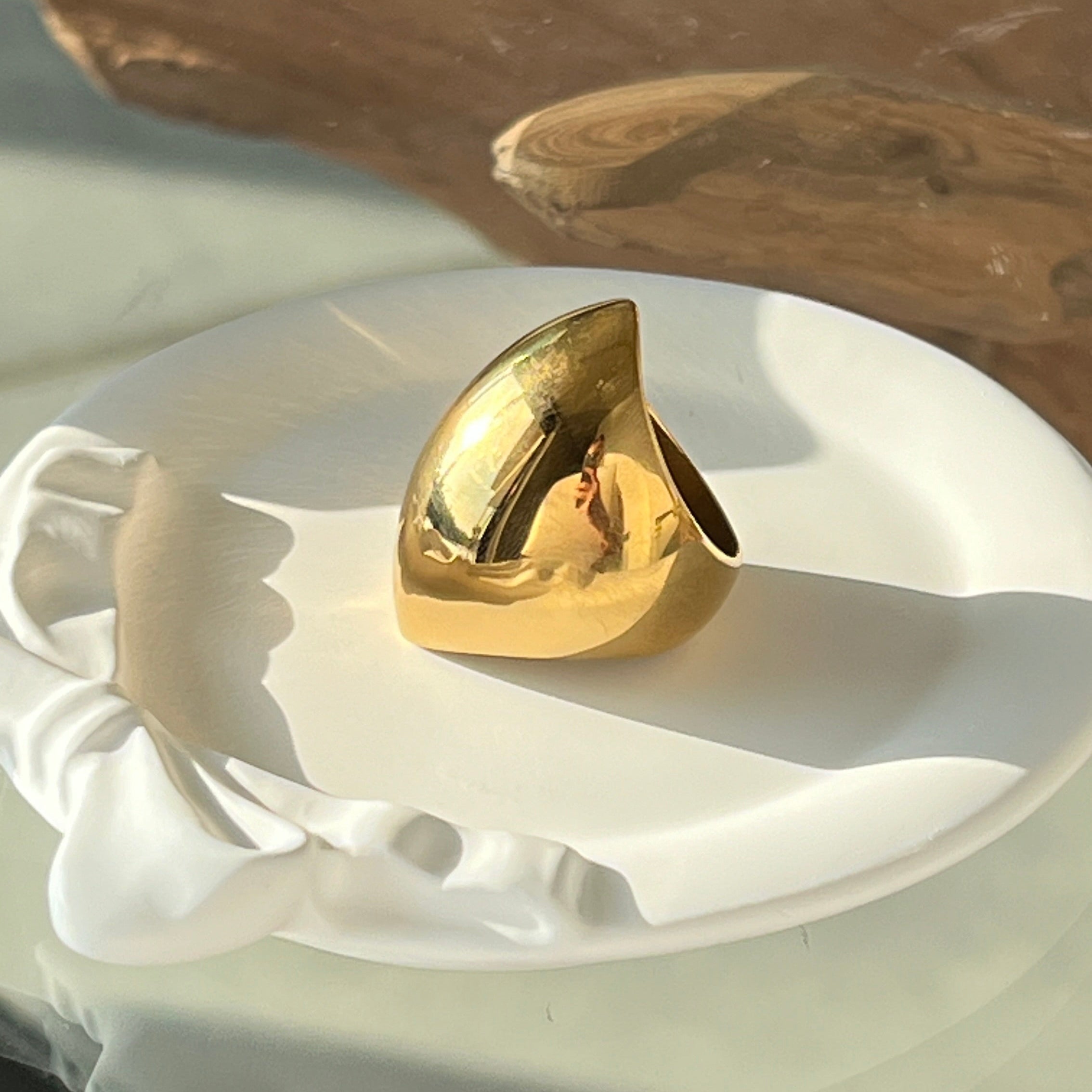 Angular ring in PVD-coated 18k gold-plated stainless steel on a white dish with a wooden background in natural light