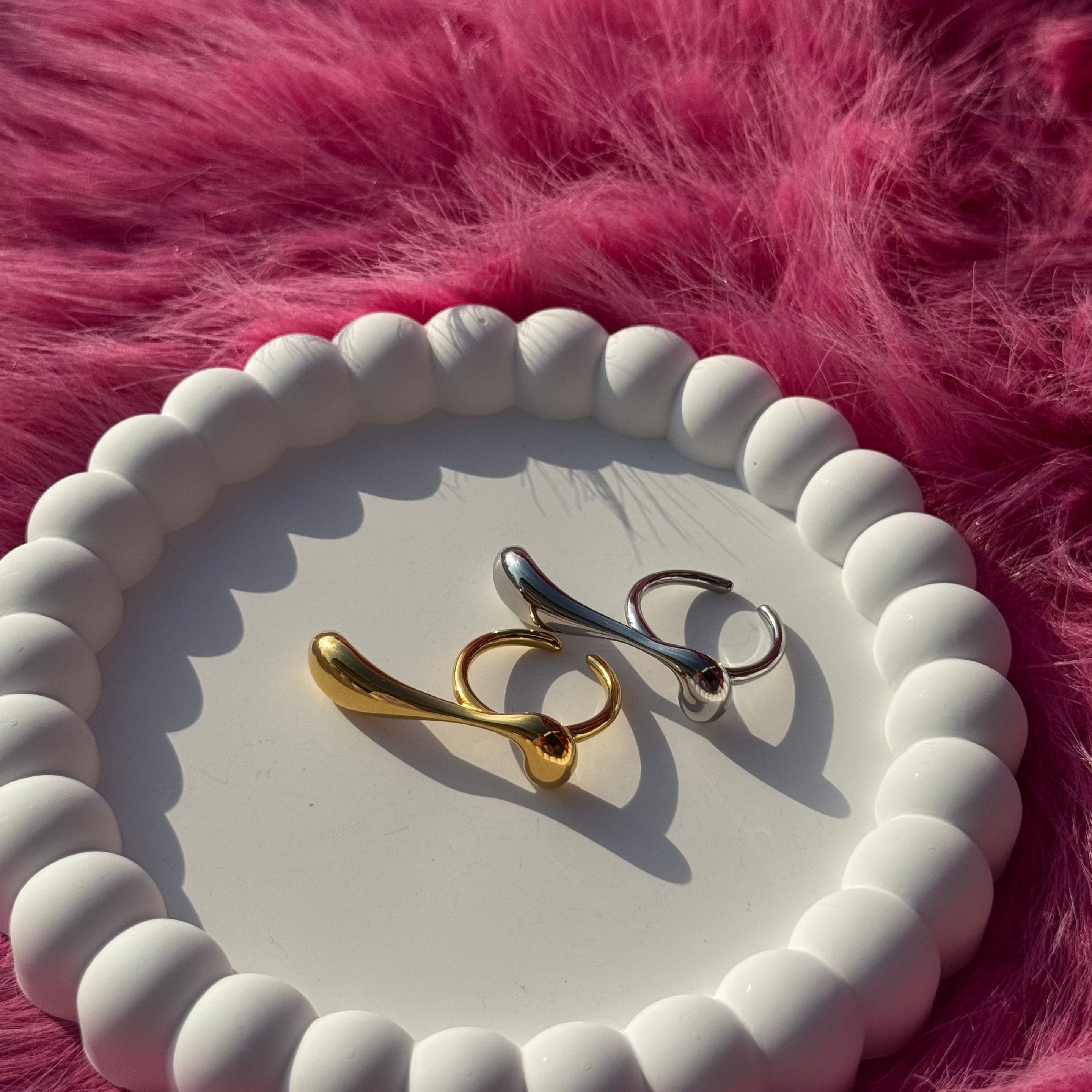 Long drop-shaped ring in PVD-coated 18k gold-plated stainless steel shown beside a silver version on a white beaded dish against a pink fur background