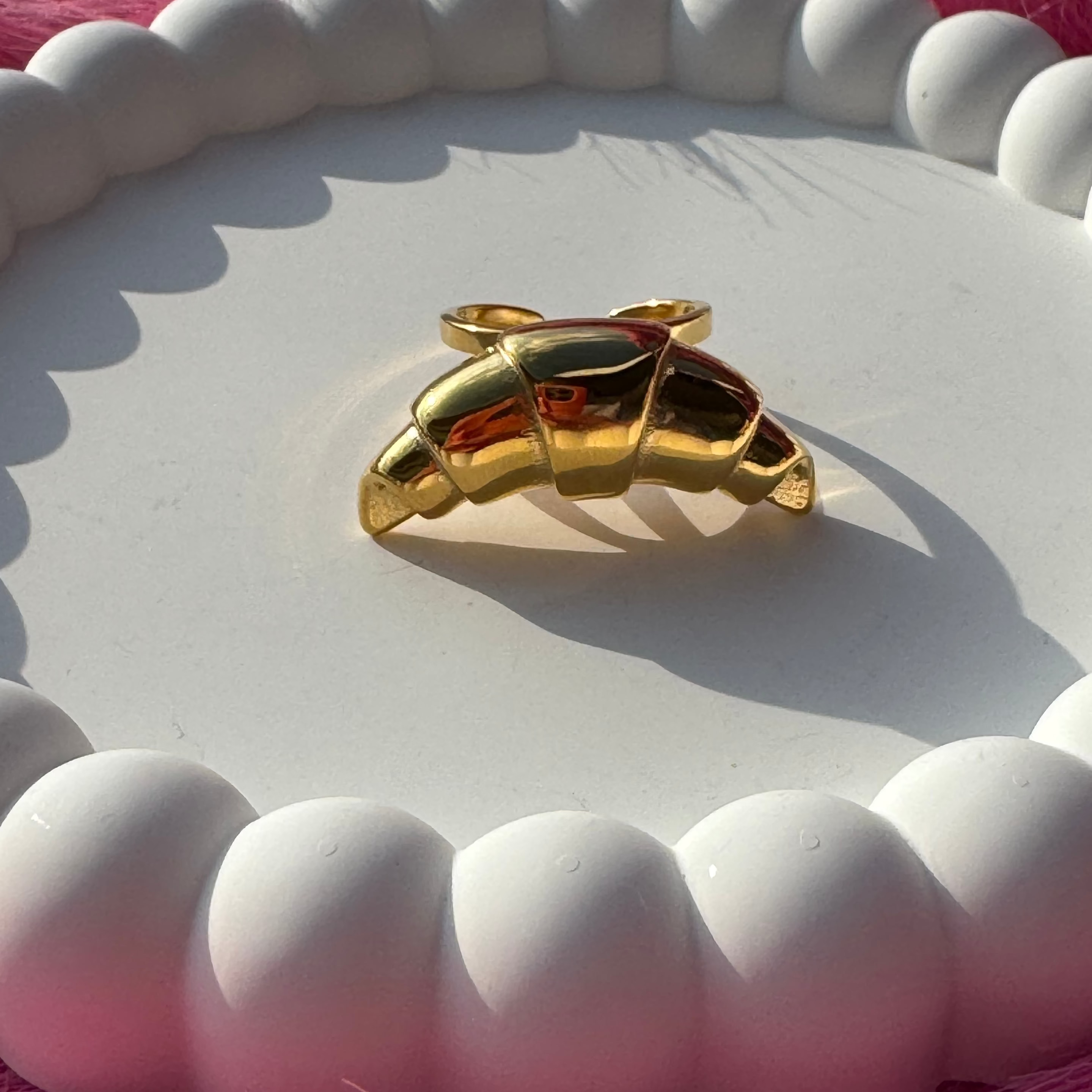 Croissant-shaped drop ring in PVD-coated 18k gold-plated stainless steel on a white beaded dish against a pink fur background