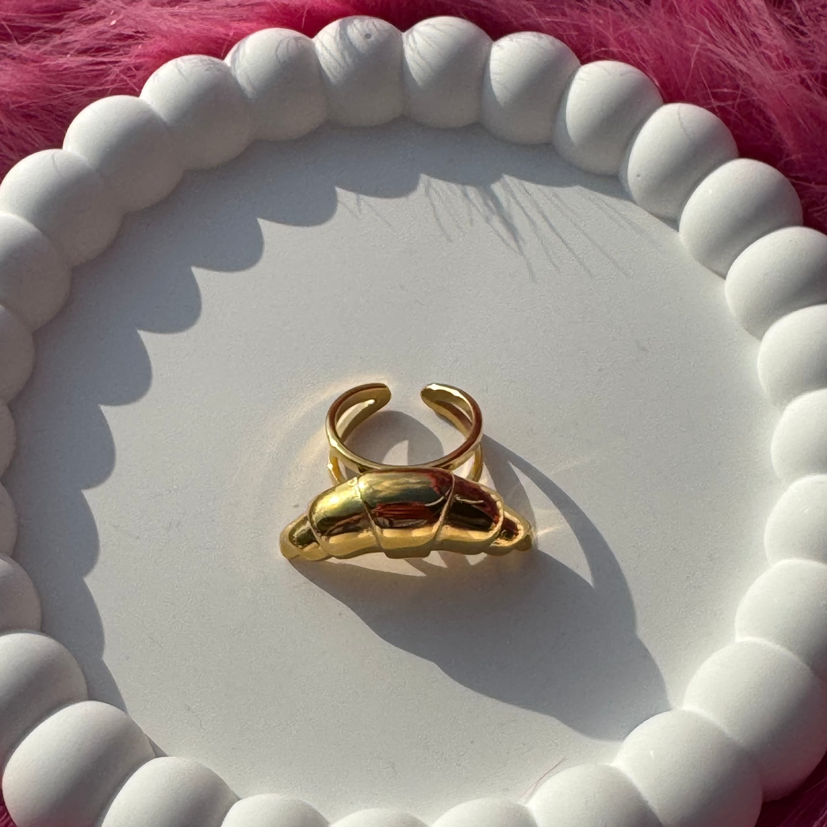 Top view of the croissant-shaped dome ring in PVD-coated 18k gold-plated stainless steel on a white beaded dish against a pink fur background