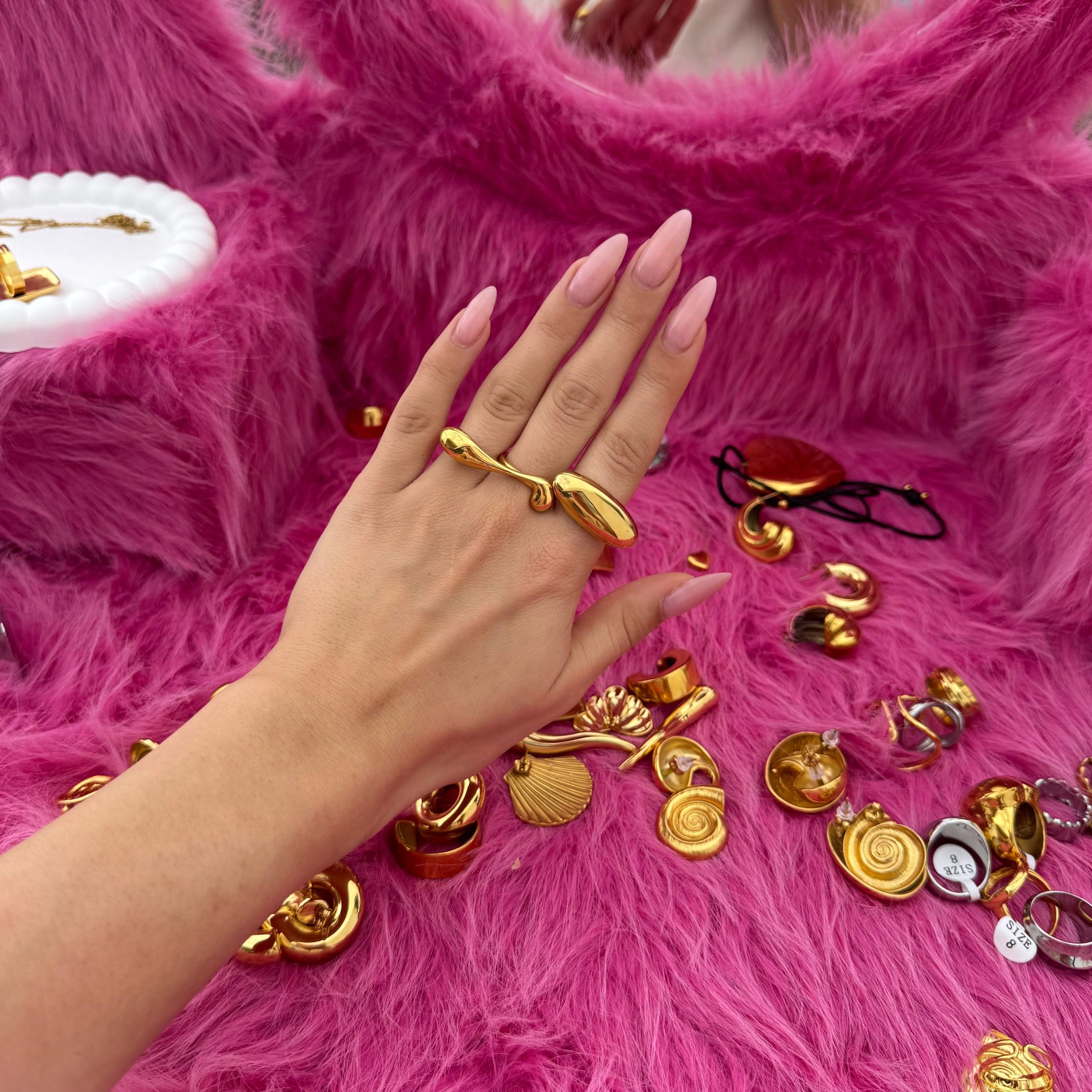 Hand wearing the long drop-shaped ring in PVD-coated 18k gold-plated stainless steel resting on pink fur scattered with gold jewellery