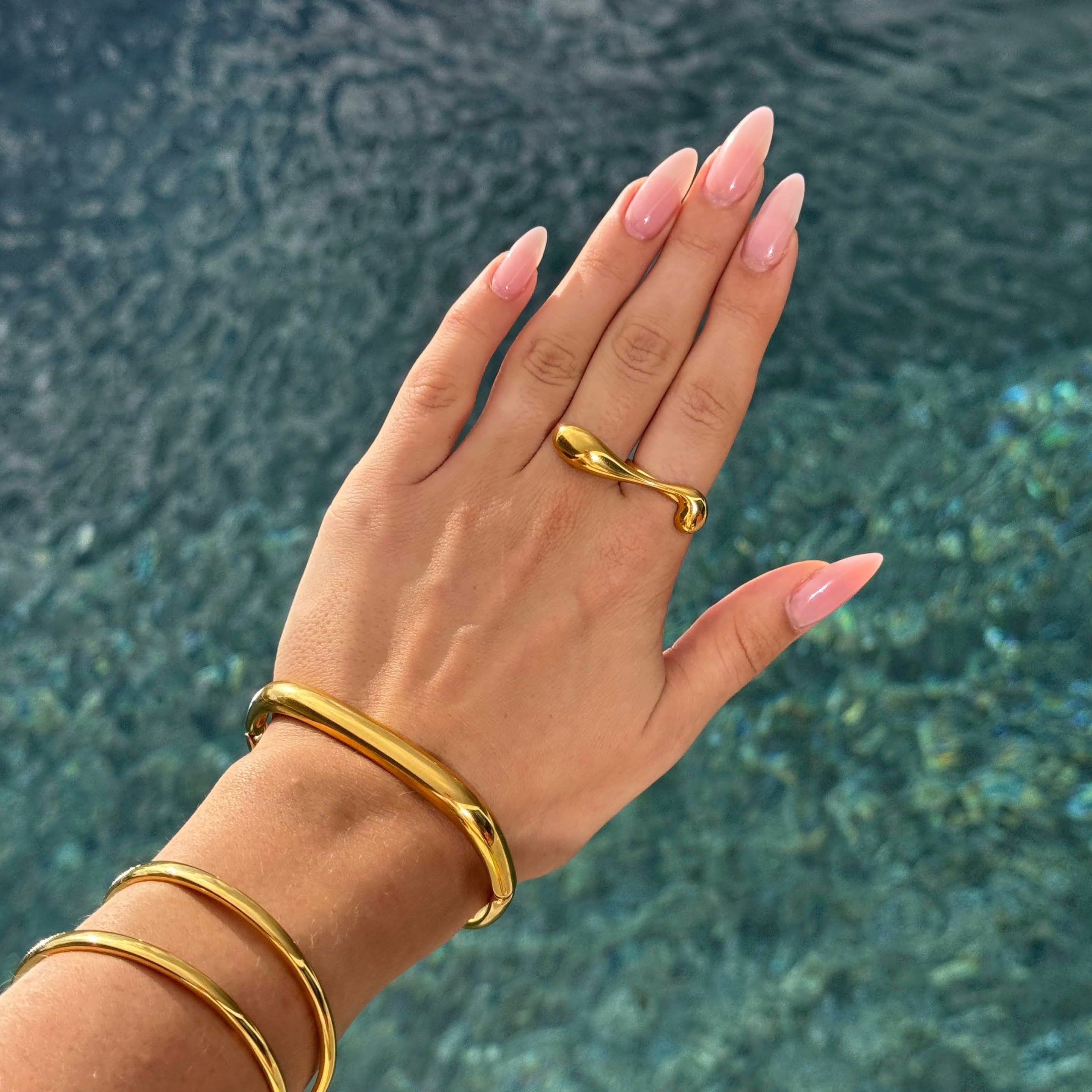 Hand wearing the long drop-shaped ring in PVD-coated 18k gold-plated stainless steel with matching gold cuffs, held over sunlit water to show its waterproof finish