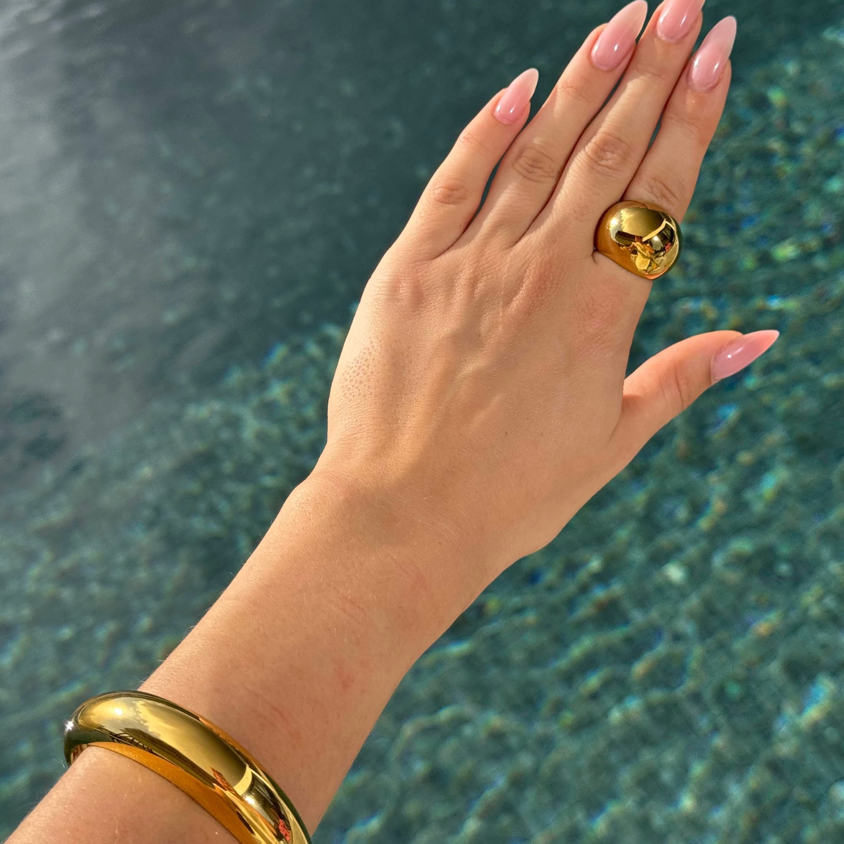 Hand wearing the ball ring in PVD-coated 18k gold-plated stainless steel with a matching gold cuff, held over sunlit water to show its waterproof finish