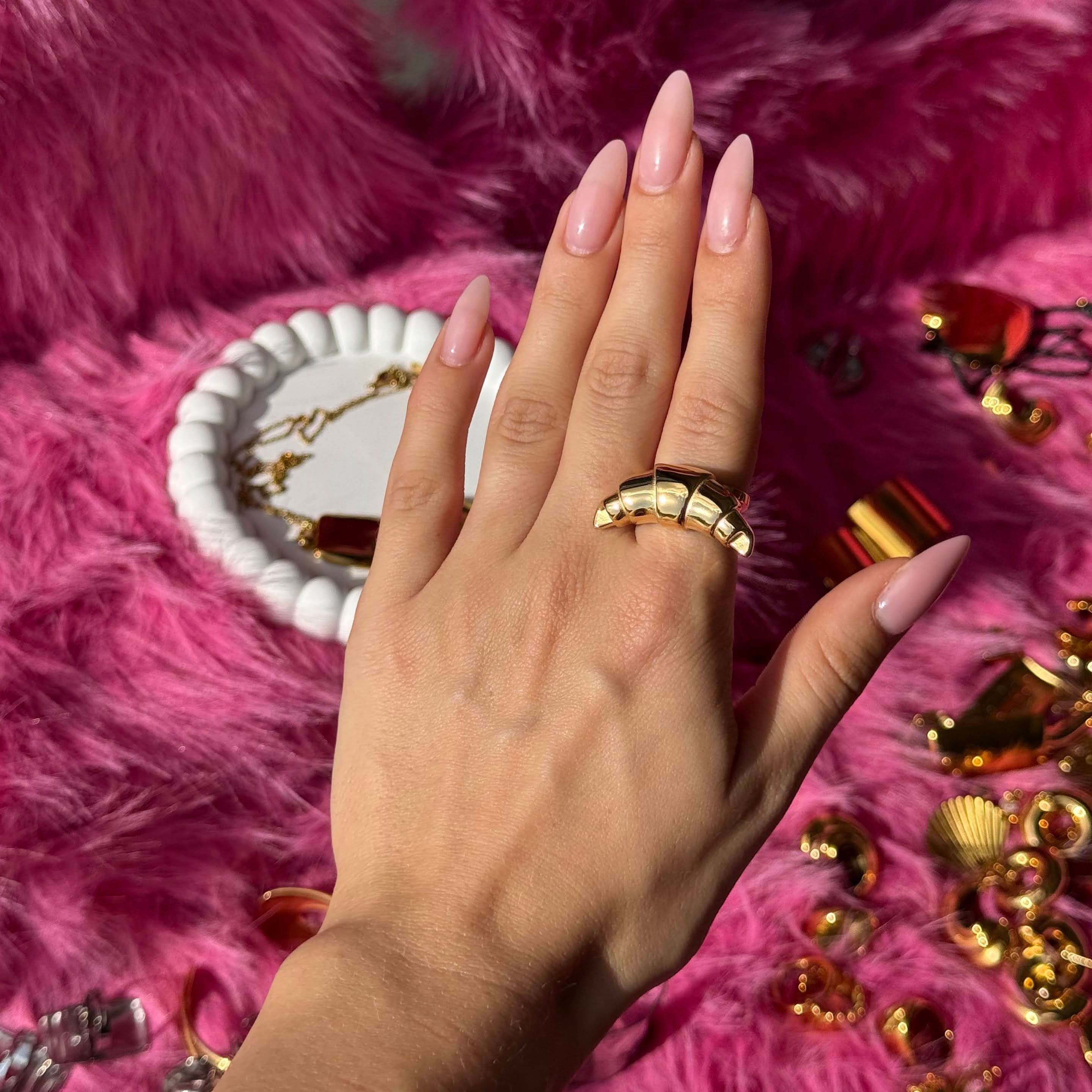Hand wearing the croissant-shaped dome ring in PVD-coated 18k gold-plated stainless steel, surrounded by gold jewellery on a pink fur background