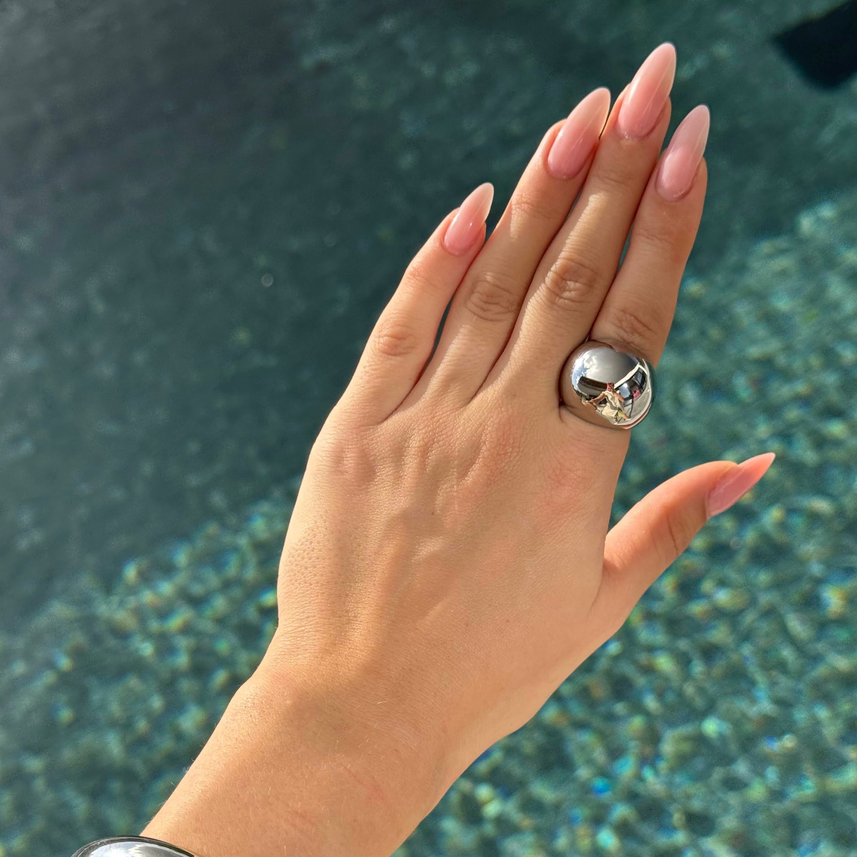 Hand wearing the ball ring in PVD-coated 925 silver-plated stainless steel with a matching silver cuff, held over sunlit water to show its waterproof finish