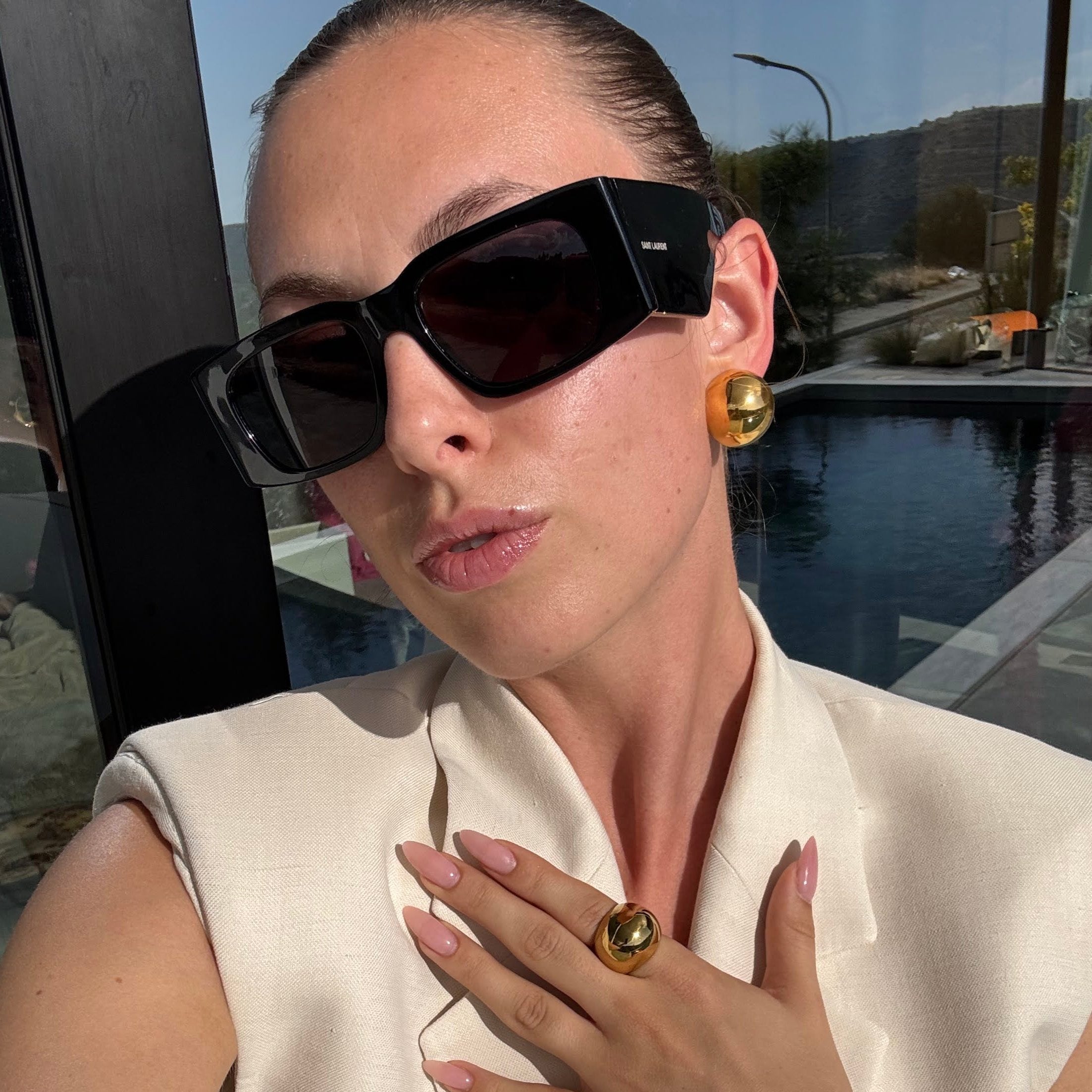 Model in sunglasses and a cream blazer wearing the ball ring in PVD-coated 18k gold-plated stainless steel with matching gold earrings and cuff