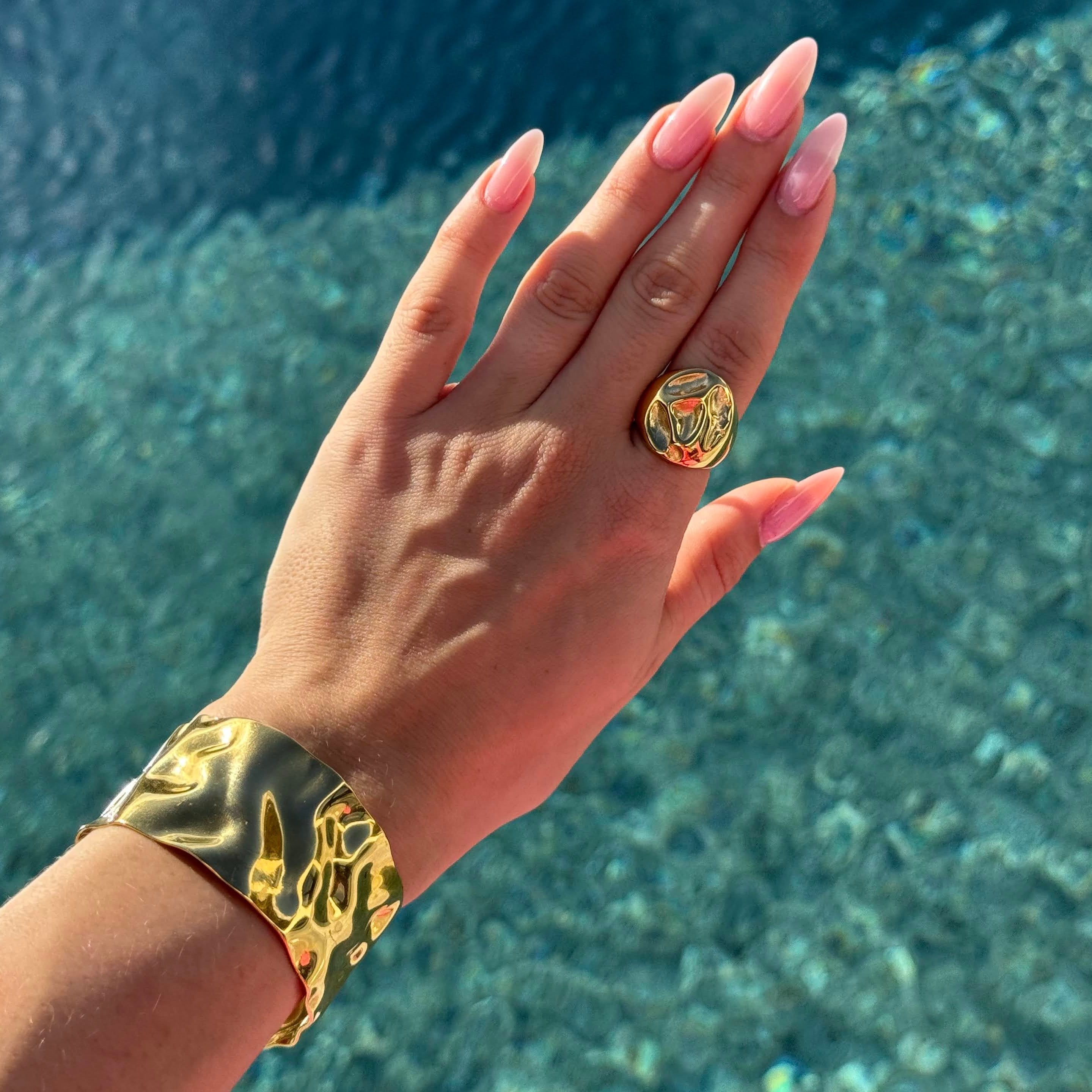 Hand wearing the irregular bold ring in PVD-coated 18k gold-plated stainless steel and a gold cuff against a blue water background