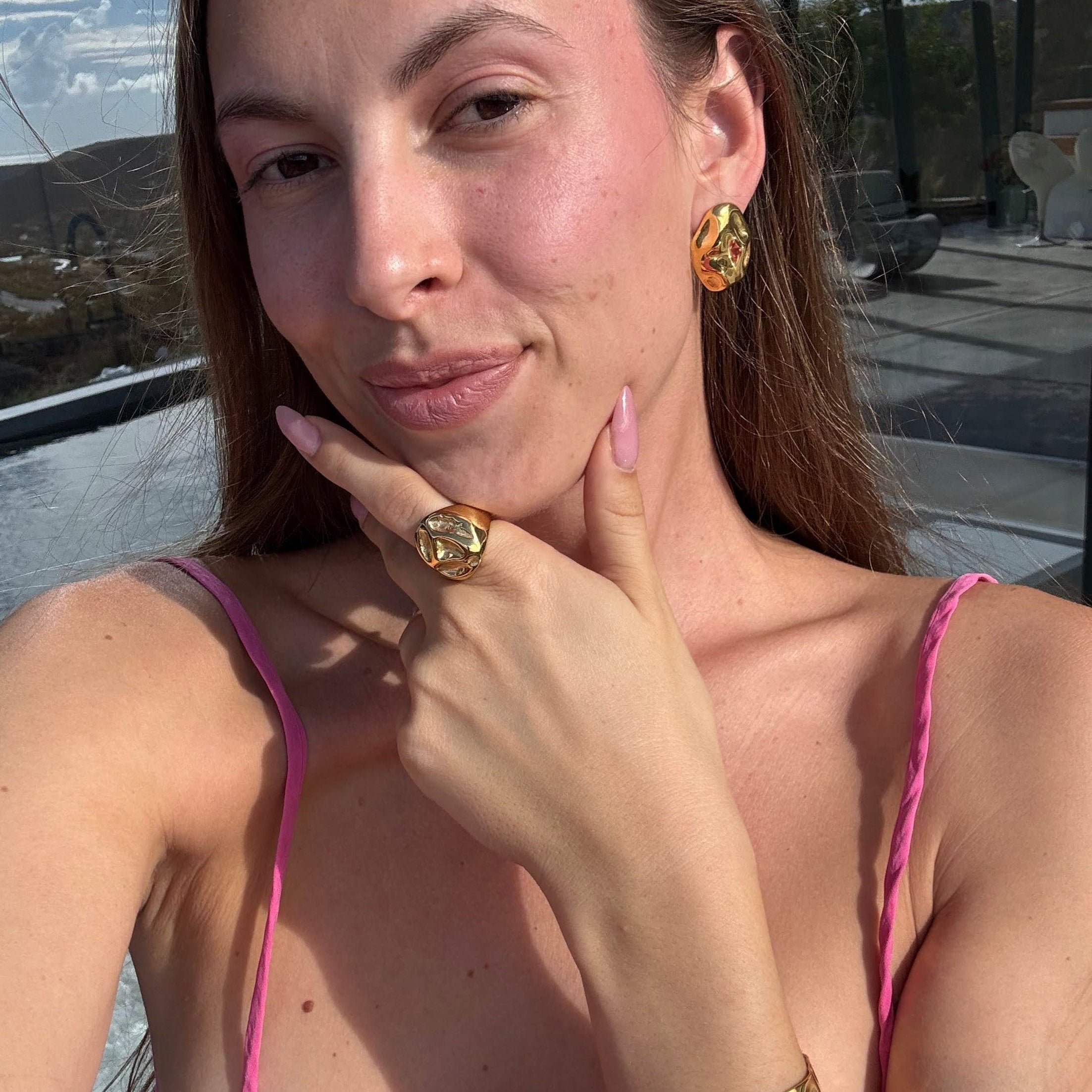 Model in a pink top wearing the irregular bold ring in PVD-coated 18k gold-plated stainless steel with matching gold jewellery