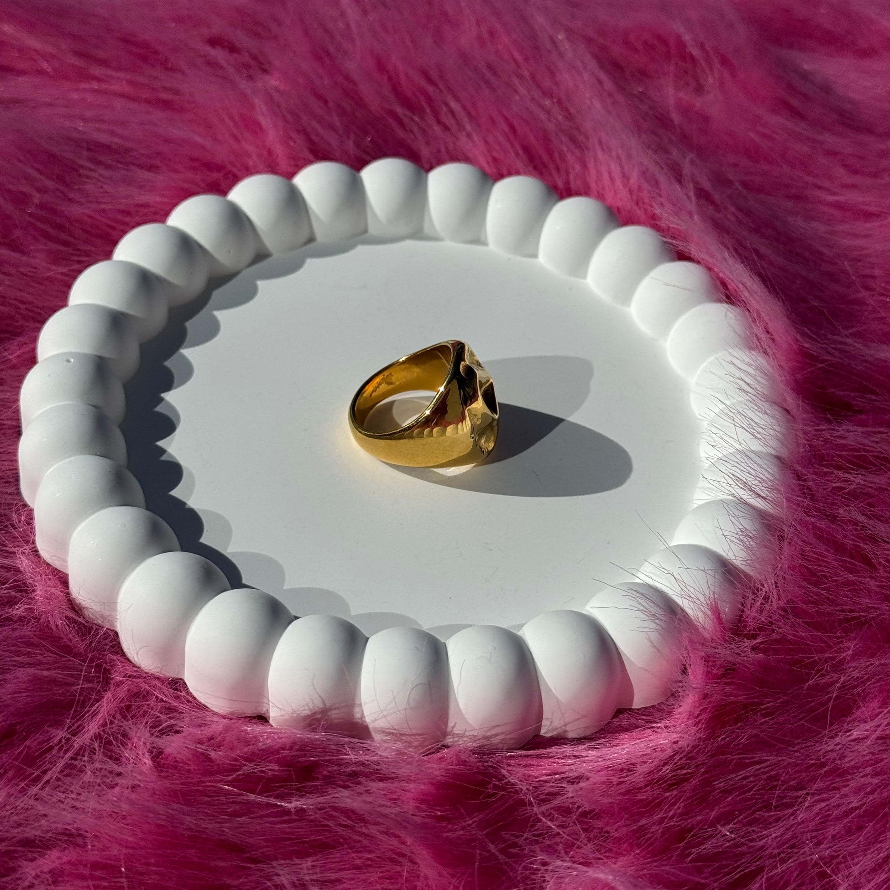 Side view of the irregular bold chunky ring in PVD-coated 18k gold-plated stainless steel on a white beaded dish against a pink fur background