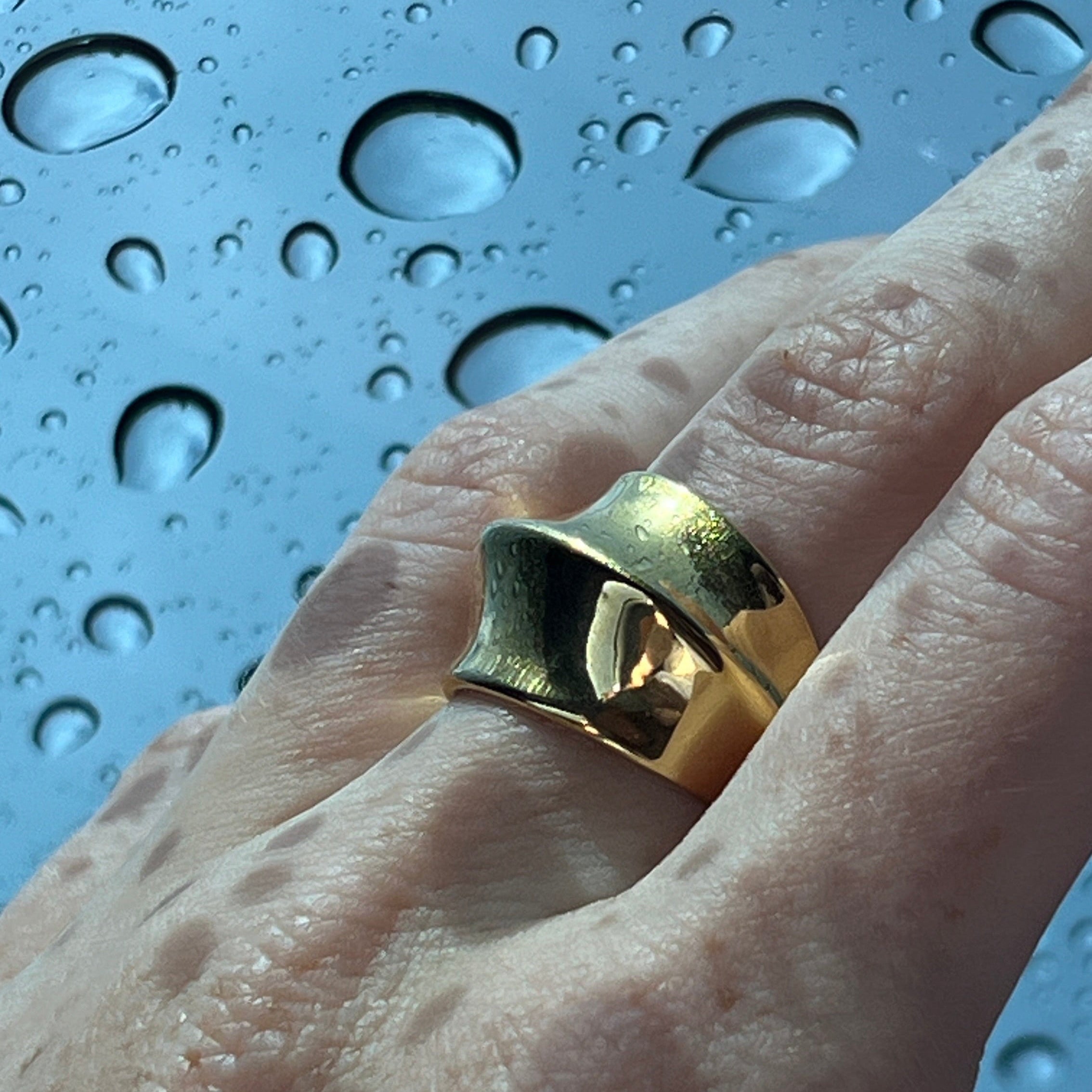 Close-up of the fashionable dome ring in PVD-coated 18k gold-plated stainless steel on a finger against a backdrop of water droplets, showing its waterproof finish