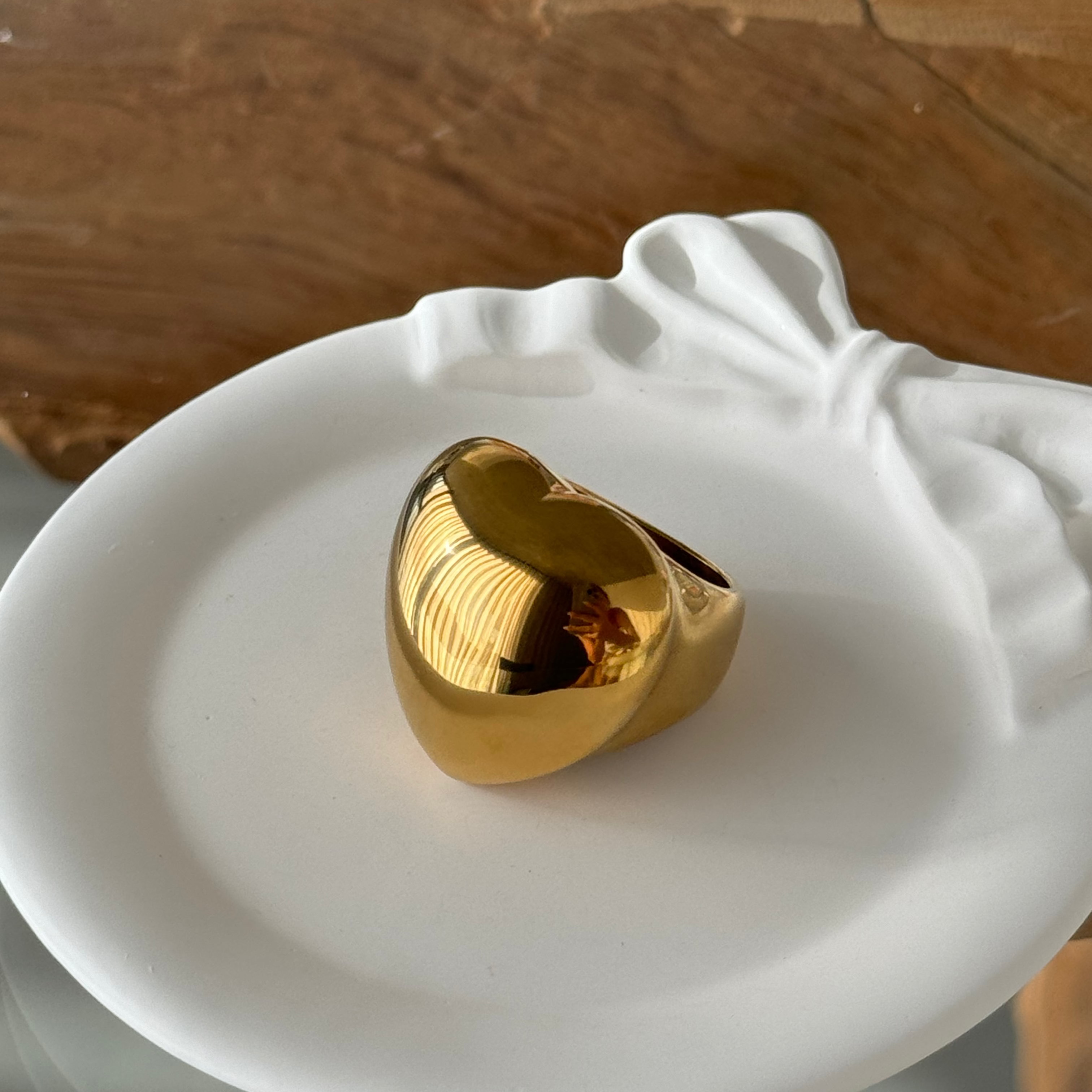 Love-shaped ring in PVD-coated 18k gold-plated stainless steel resting on a white ceramic dish, its smooth domed heart silhouette catching the light; a waterproof, tarnish-free band