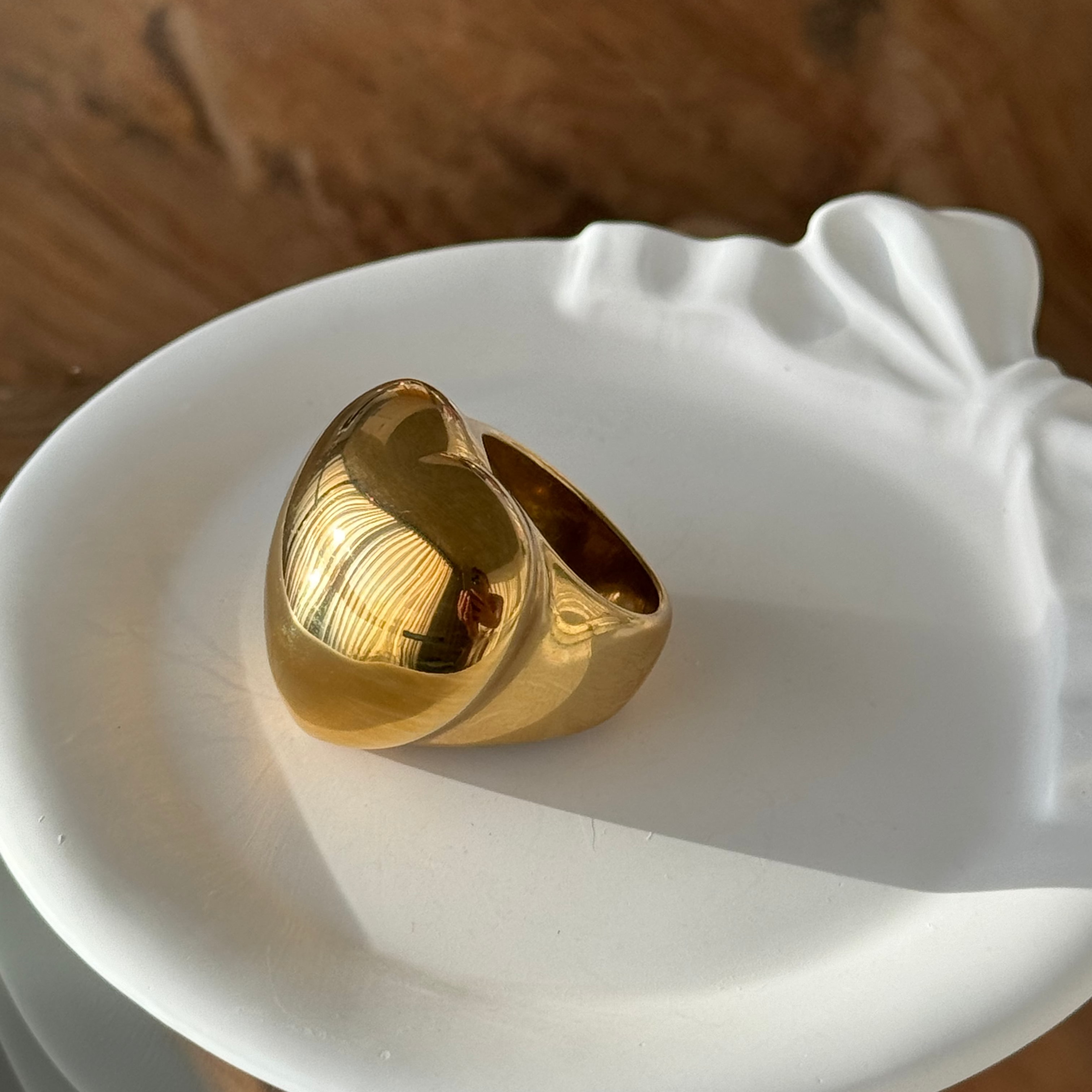 Side profile of the love-shaped ring in PVD-coated 18k gold-plated stainless steel on a white dish in warm sunlight, showing its chunky domed heart band; waterproof and tarnish-free