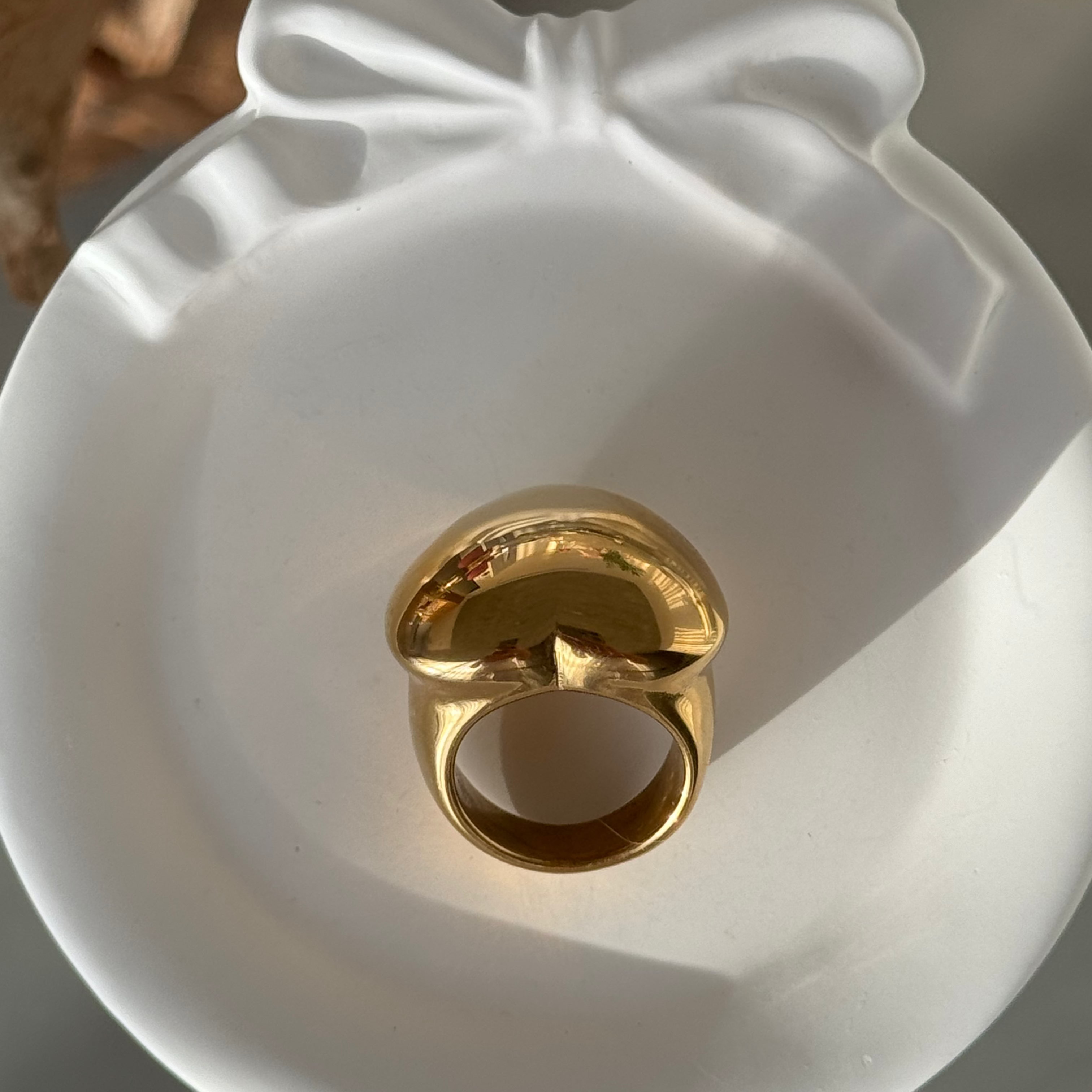 Top-down view of the love-shaped ring in PVD-coated 18k gold-plated stainless steel in a white bow-shaped dish, showing its rounded heart face and open band; a waterproof, hypoallergenic ring