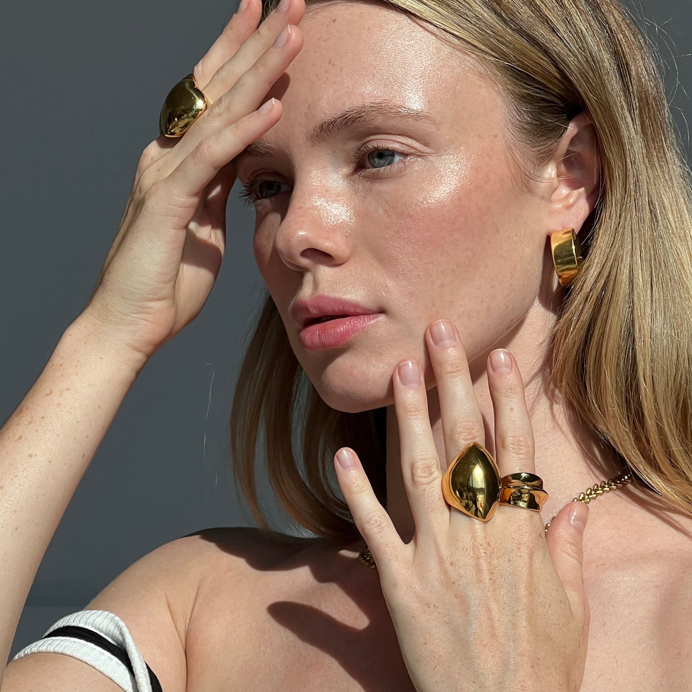 Blonde model wearing the angular ring in PVD-coated 18k gold-plated stainless steel and matching gold jewellery with hands framing her face