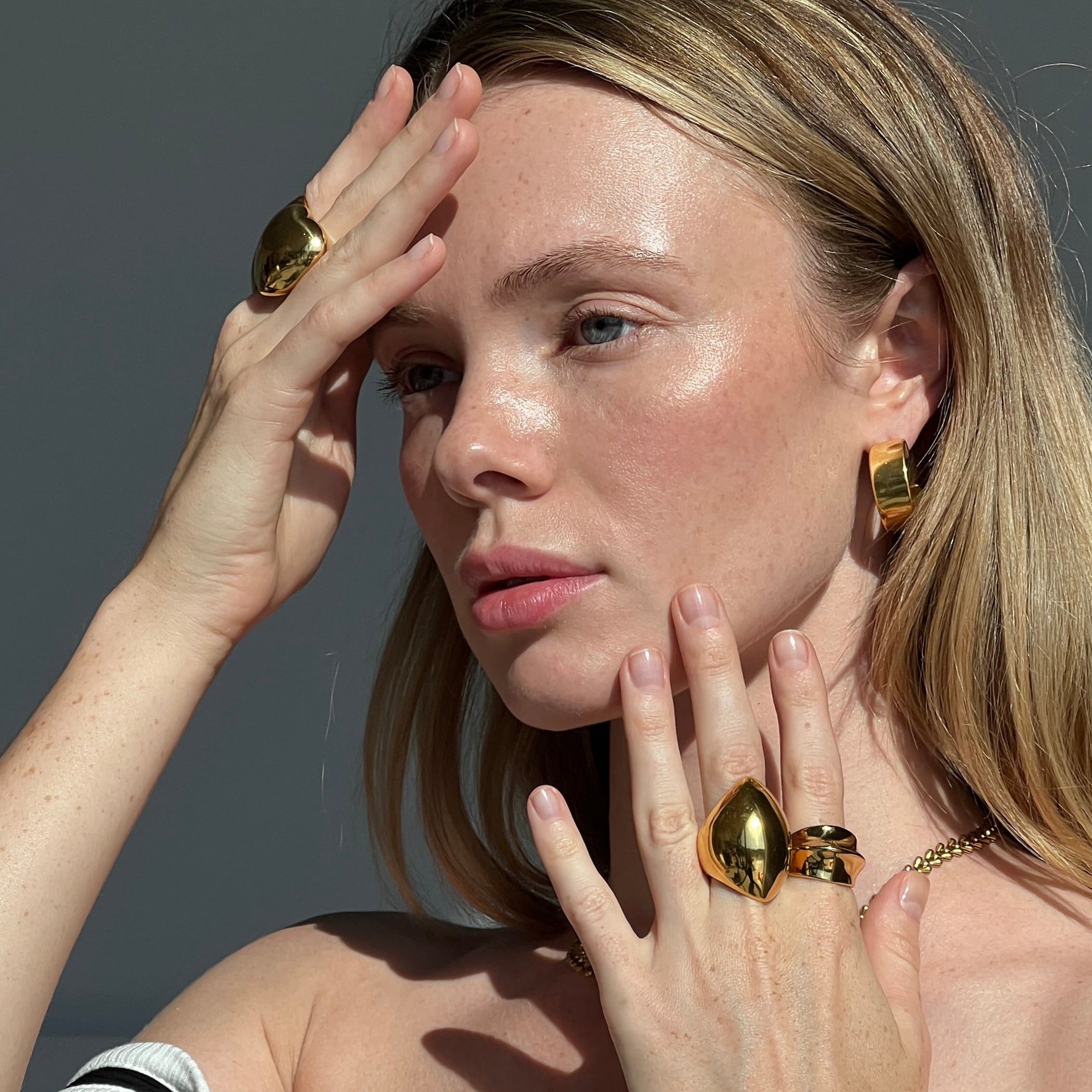 Blonde model wearing the fashionable dome ring in PVD-coated 18k gold-plated stainless steel with matching gold jewellery, hands framing her face