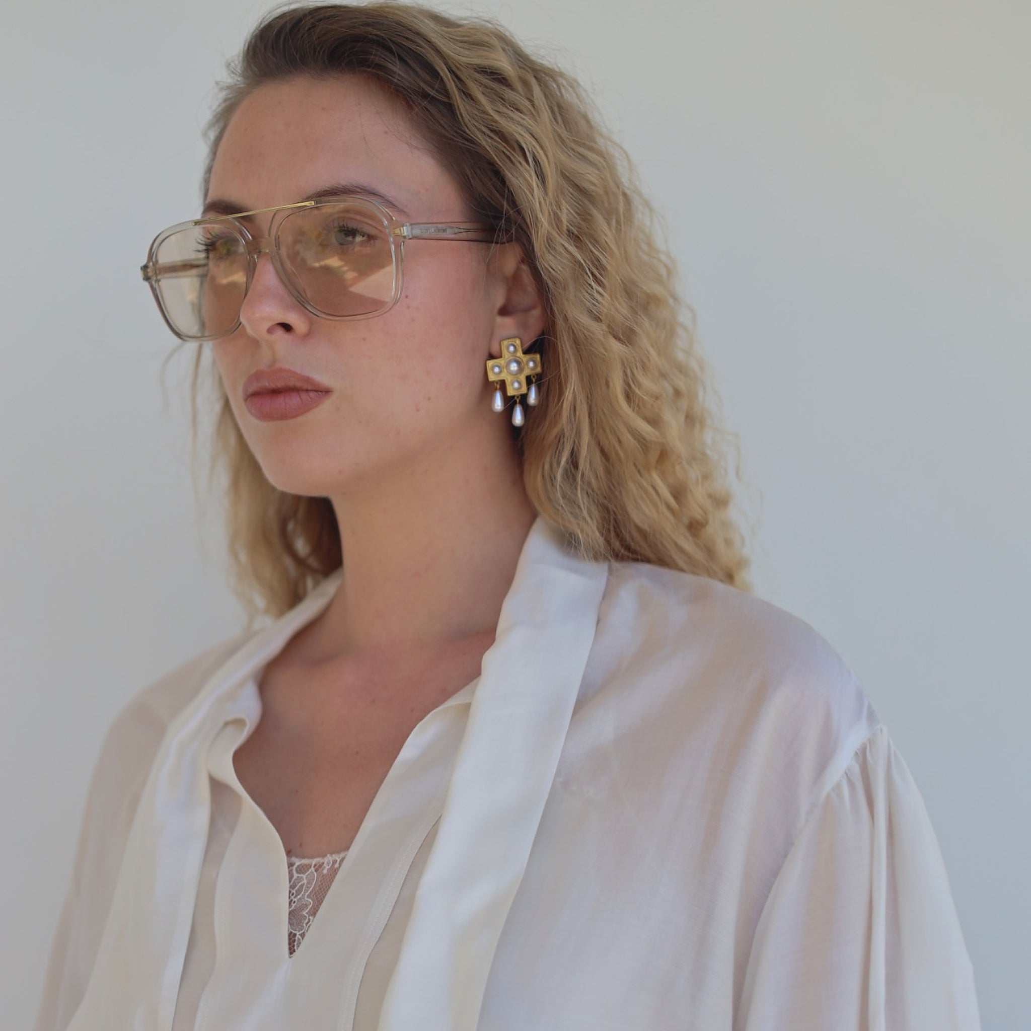 Blonde model in sunglasses and a white blouse wearing the Pearls Before Drama gold cross drop earring with faux-pearl drops