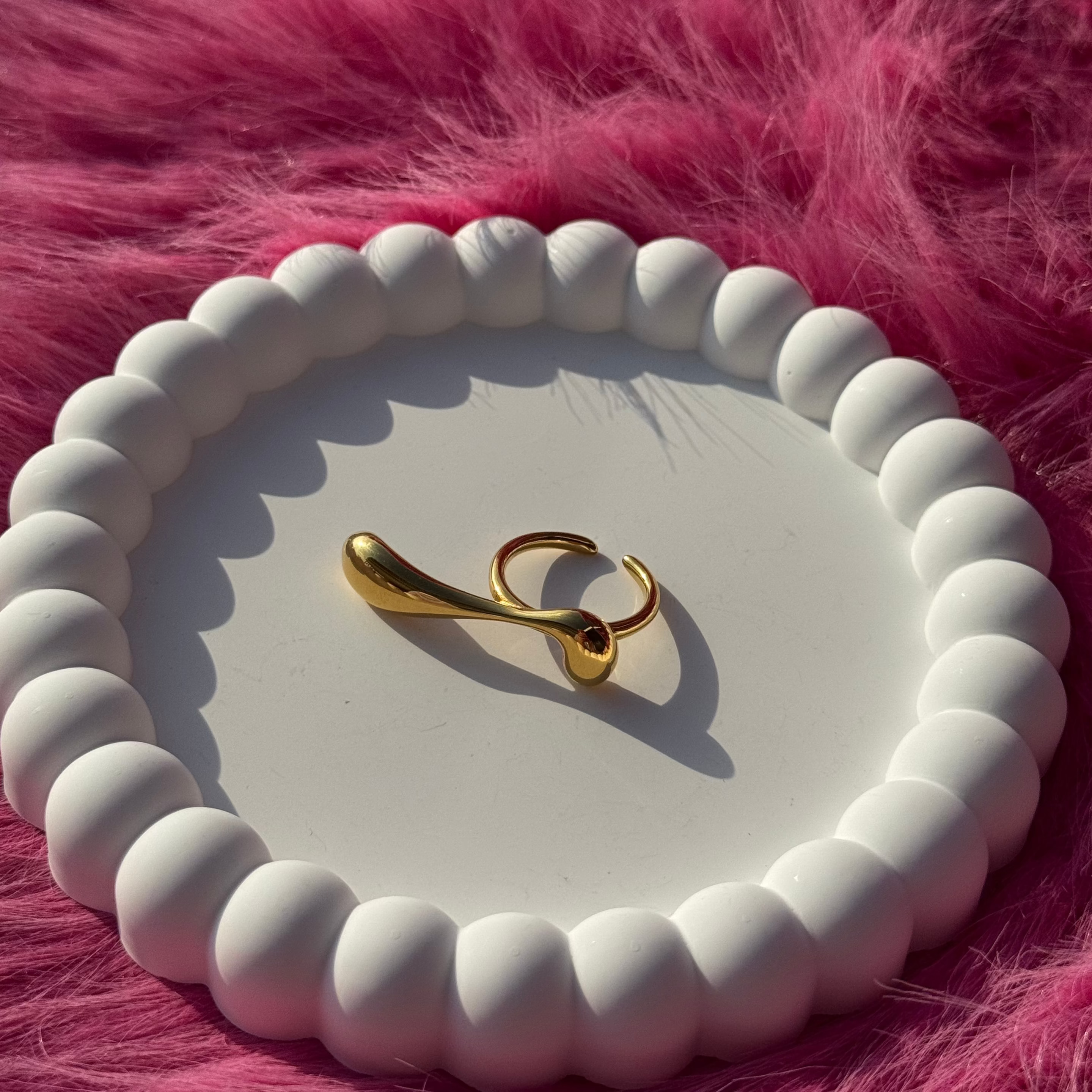 Long drop-shaped ring in PVD-coated 18k gold-plated stainless steel displayed on a white beaded dish against a pink fur background