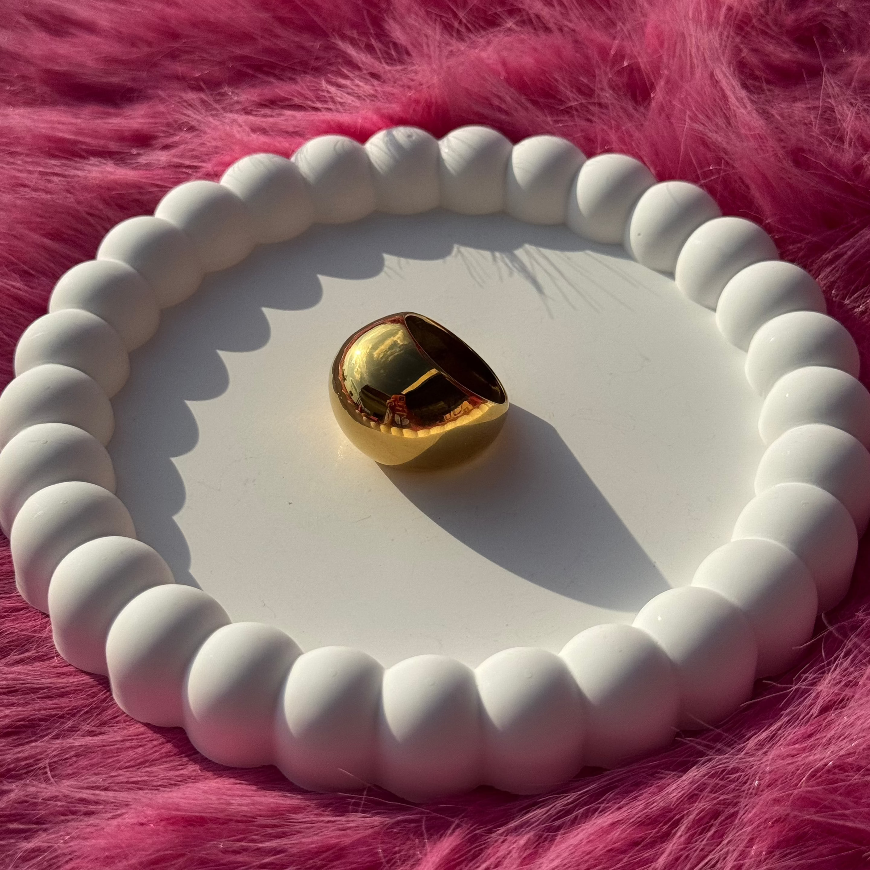 Single ball ring in PVD-coated 18k gold-plated stainless steel displayed on a white beaded dish against a pink fur background