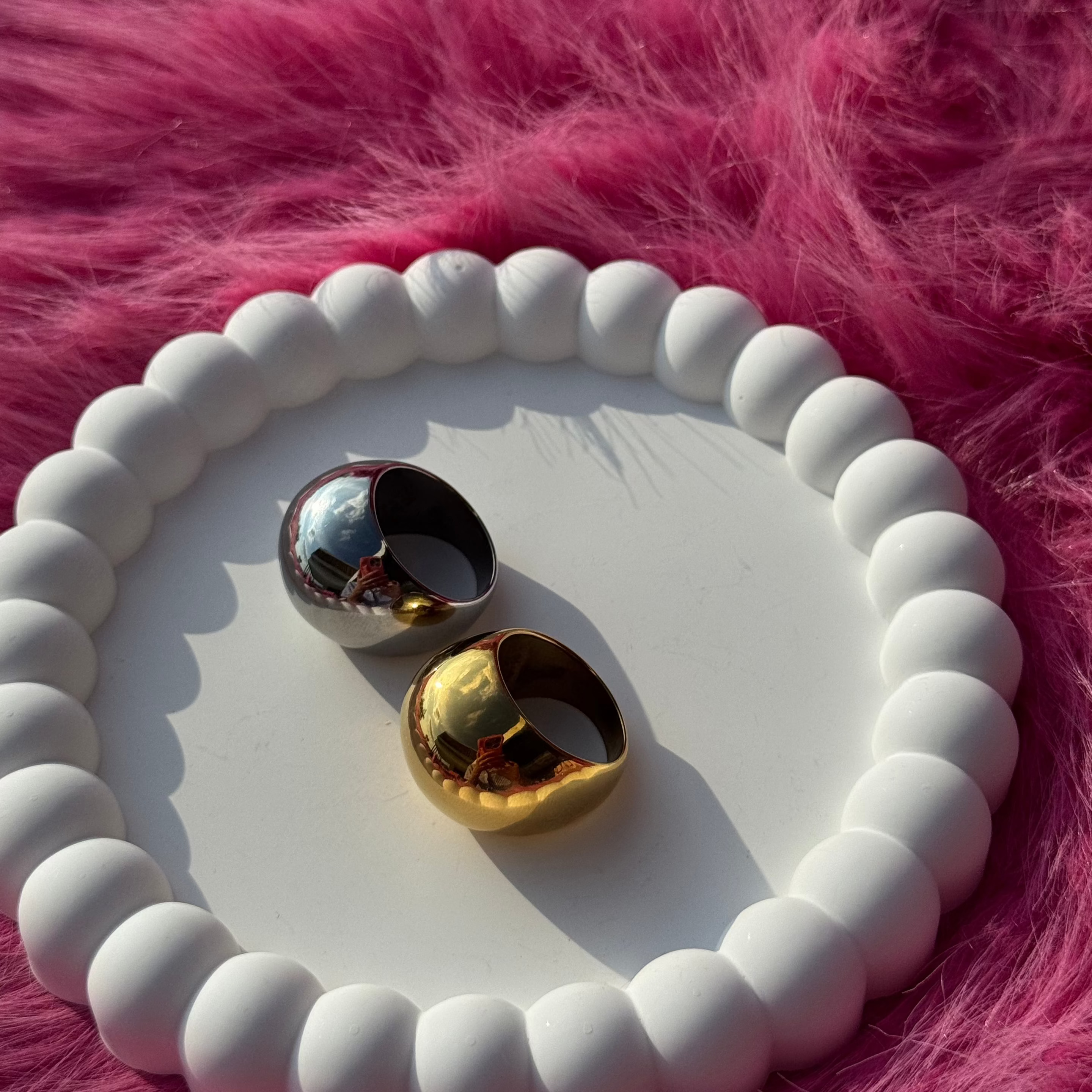 Ball ring in PVD-coated 925 silver-plated stainless steel shown beside a gold version on a white beaded dish against a pink fur background