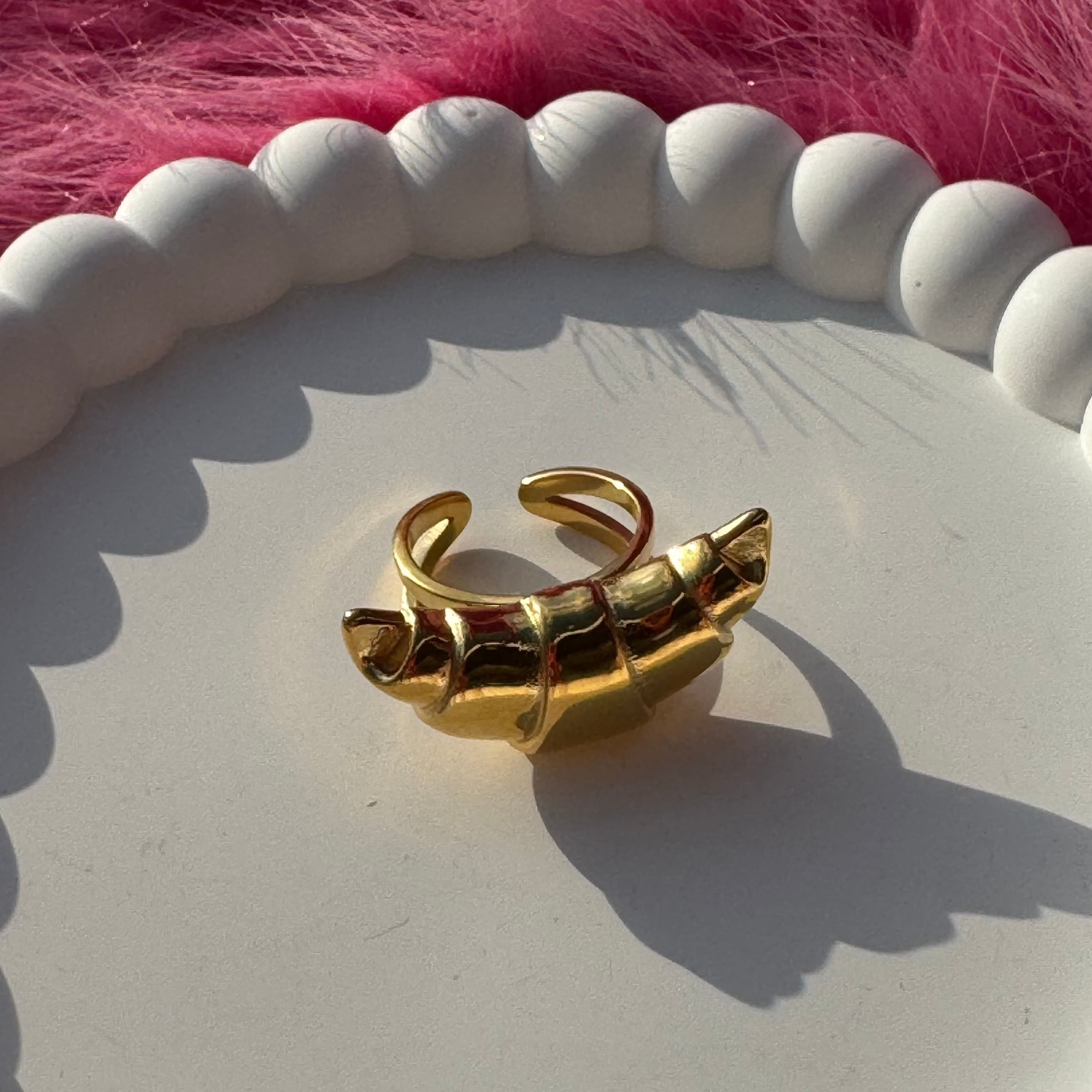 Side view of the croissant-shaped open ring in PVD-coated 18k gold-plated stainless steel on a white beaded dish against a pink fur background