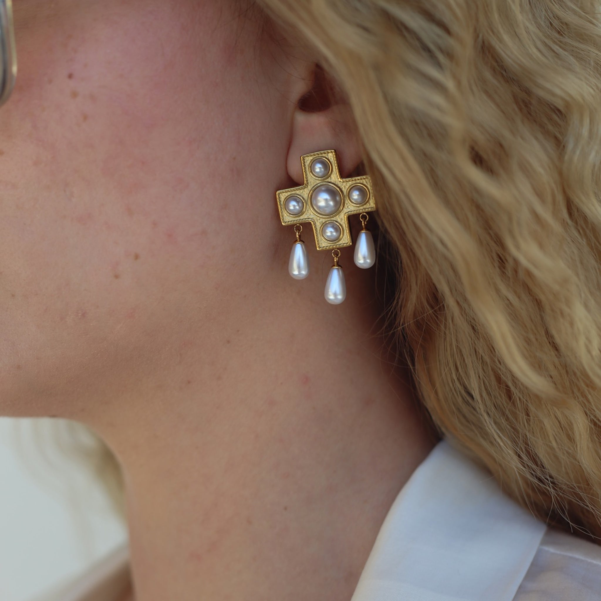 Close-up of a model in a white blouse wearing the Pearls Before Drama gold cross drop earring with cubic zirconia and faux-pearl drops